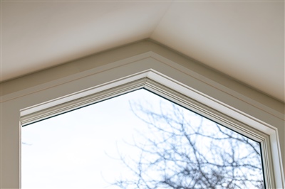 Close-up view of a house interior showing a detailed white crown molding around a triangular window, framing a view of bare tree branches against a gray sky.