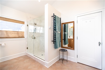 Minimalist bathroom with a glass shower enclosure, white walls, a wooden shelf, and a small mirrored cabinet beside the entry door.