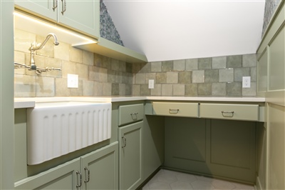 A compact utility room with green cabinets, tiled backsplash, and under-cabinet lighting, featuring a large sink and radiator.