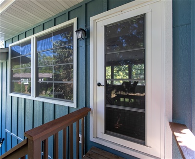 Exterior view of a house entry with teal siding, featuring white windows and a dark door, set on a wooden porch.