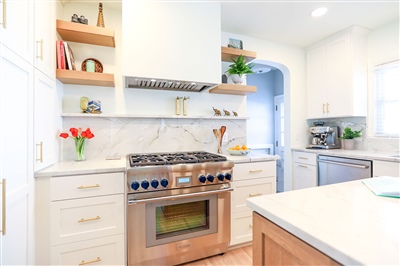 Bright kitchen with white cabinetry, marble countertops, and stainless steel appliances, complemented by wooden open shelving and decorative plants.