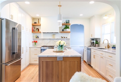 Bright kitchen with white cabinetry, marble countertops, and stainless steel appliances, complemented by wooden open shelving and decorative plants.