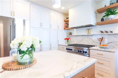 Bright kitchen with white cabinetry, marble countertops, and stainless steel appliances, complemented by wooden open shelving and decorative plants.