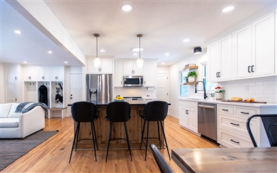 Open concept kitchen with a central breakfast bar, black stools, white cabinets, and wooden floors, leading to a living area.