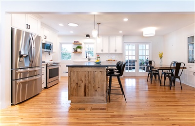 Open concept kitchen with a central breakfast bar, black stools, white cabinets, and wooden floors, leading to a living area.