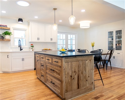 Open concept kitchen with a central breakfast bar, black stools, white cabinets, and wooden floors, leading to a living area.