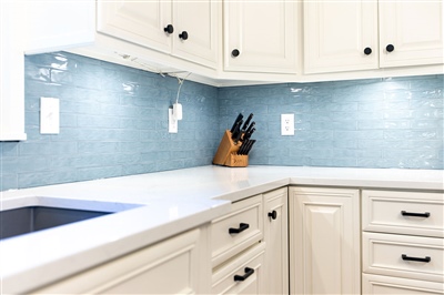 Modern kitchen corner with white cabinets, quartz countertops, and a blue tiled backsplash, highlighted by under-cabinet lighting.