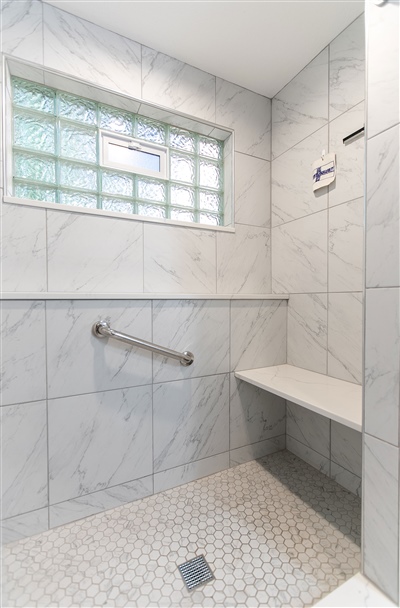 Shower area with marble tiling throughout, featuring a built-in bench, safety handle, and a window for natural light.